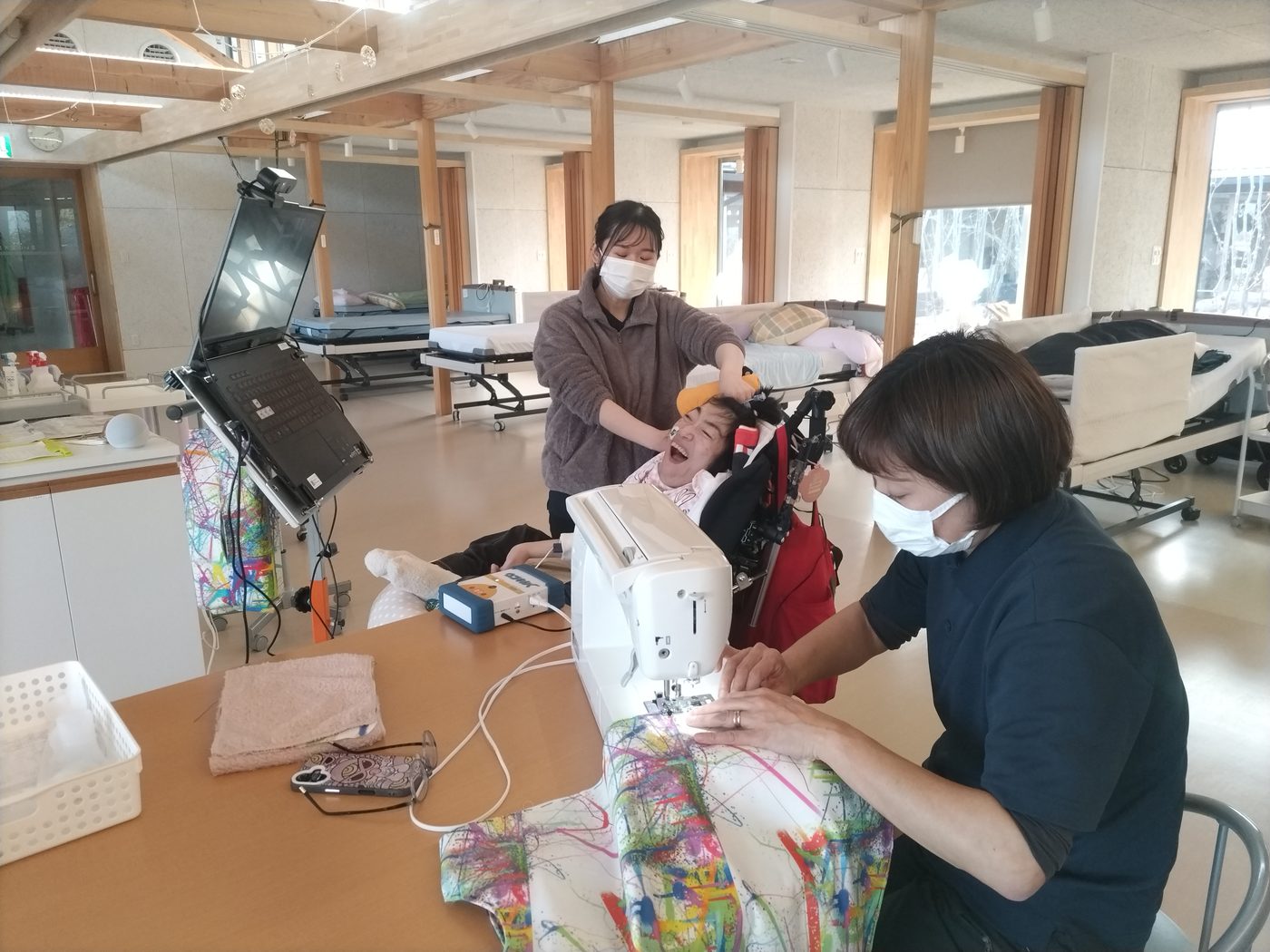 学習者が笑顔を見せ、そばで職員が布をミシンで縫っている